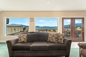 A living room with a brown sofa, two windows with a view of the sea, and a door leading outside.