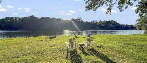A grassy area with a lake, chairs, and a table under a tree.