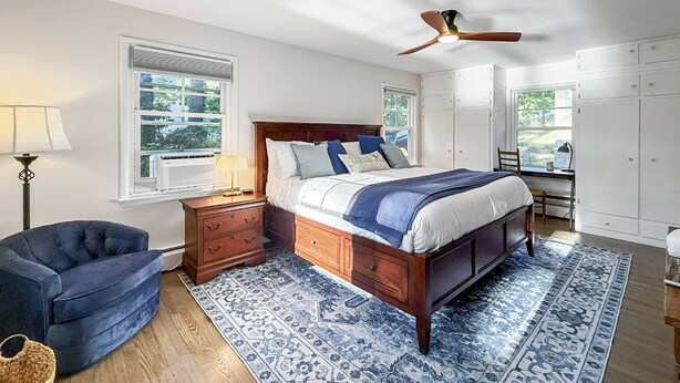 Un dormitorio con cama de madera, un sillón azul, una mesita de noche, una lámpara de pie y un ventilador de techo.