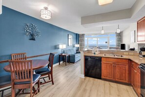 A modern kitchen with a dishwasher, a dining area with a table and chairs, a blue accent wall, and a view of the ocean. - Dunes Village Oceanfront Condo + Waterpark (Myrtle Beach)