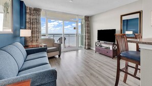 Ein Hotelzimmer mit einer Couch, einem Fernseher, einem Esstisch mit Stühlen und einem Balkon mit Meerblick.