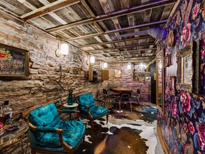 A rustic interior with stone walls, wooden ceiling beams, and a cowhide floor. There are turquoise chairs, a small round table, and a bar area with a bottle. - Historic Manor w/ Hidden Speakeasy, 3BR/2.5BA (Heber City)