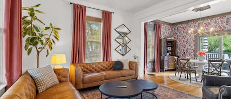 A spacious living room with a leather sofa, a round coffee table, and a patterned rug. There is a dining area with a table and chairs visible through an open doorway.