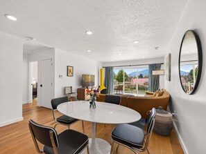A modern living room with a round dining table, black chairs, a sofa, and a large window with a view of a red-roofed building. - Swanky Mid Century Gem, 2BR/1BA, Deck, Fireplace (Heber City)