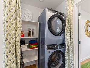 A stacked front-loading washer and dryer in a laundry room with shelves above containing folded laundry and storage bins. - Swanky Mid Century Gem, 2BR/1BA, Deck, Fireplace (Heber City)