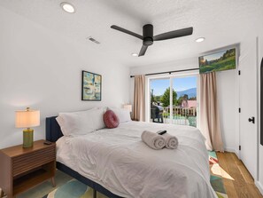 A hotel room with a bed, a nightstand, a lamp, a ceiling fan, a painting, and a view of the outdoors. - Swanky Mid Century Gem, 2BR/1BA, Deck, Fireplace (Heber City)