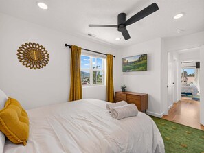 A bedroom with a bed, a ceiling fan, a window with curtains, a wall clock, a flat-screen TV, and a view of the outdoors. - Swanky Mid Century Gem, 2BR/1BA, Deck, Fireplace (Heber City)