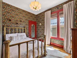 Ein Schlafzimmer mit blumigem Wandpapier, einem hölzernen Bett mit Messing-Kopfstück, einer roten Tür und einem Fenster mit Vorhängen.