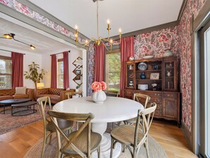 Un coin repas avec une table ronde, des chaises en bois, un lustre et un mur recouvert d’un papier peint à motifs floraux.