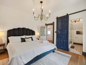 Une chambre à coucher avec un grand lit, un lustre, une porte coulissante et une salle de bain visible à travers une porte ouverte.