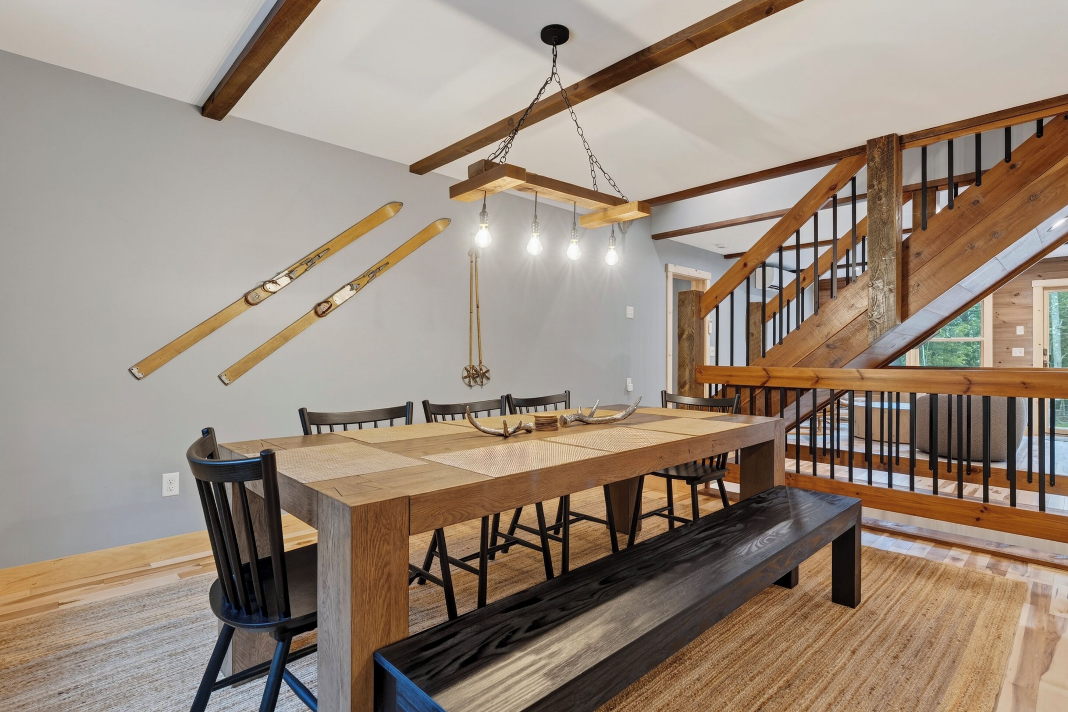 A dining area with a wooden table, chairs, and a bench. There is a staircase with wooden railings and a view of the outdoors.