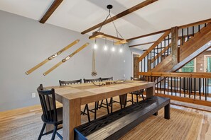 A dining area with a wooden table, chairs, and a bench. There is a staircase with wooden railings and a view of the outdoors. - New Bethel Retreat | Hot Tub, Pinball & Fireplace (Bethel)