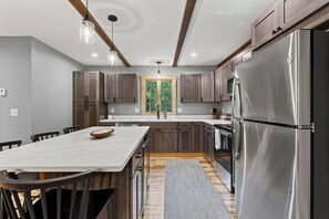 A modern kitchen with wooden cabinets, a stainless steel refrigerator, and a central island with chairs. - New Bethel Retreat | Hot Tub, Pinball & Fireplace (Bethel)