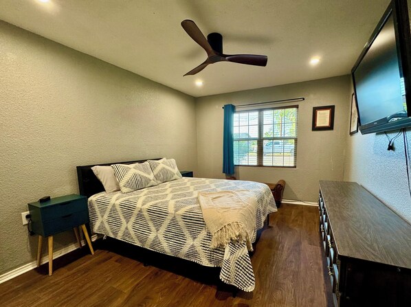 A bedroom with a bed, a nightstand, a ceiling fan, a window with curtains, and a TV mounted on the wall.