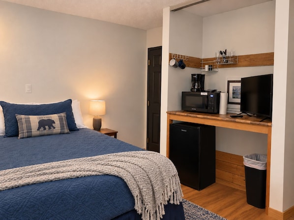 A bedroom with a bed, bedside lamp, a small desk with a coffee maker, a television, and a trash bin.