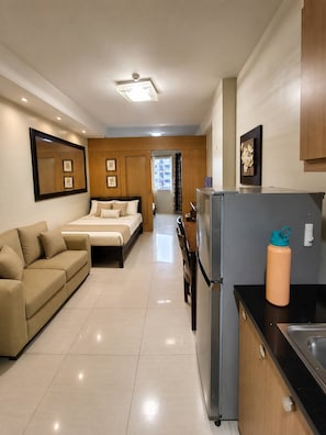 A modern hotel room with a bed, a sofa, a kitchen area with a refrigerator, and a bathroom visible through an open door. - Modern 2-bedroom Apartment With Shared Pool in Central Pasay (Pasay)