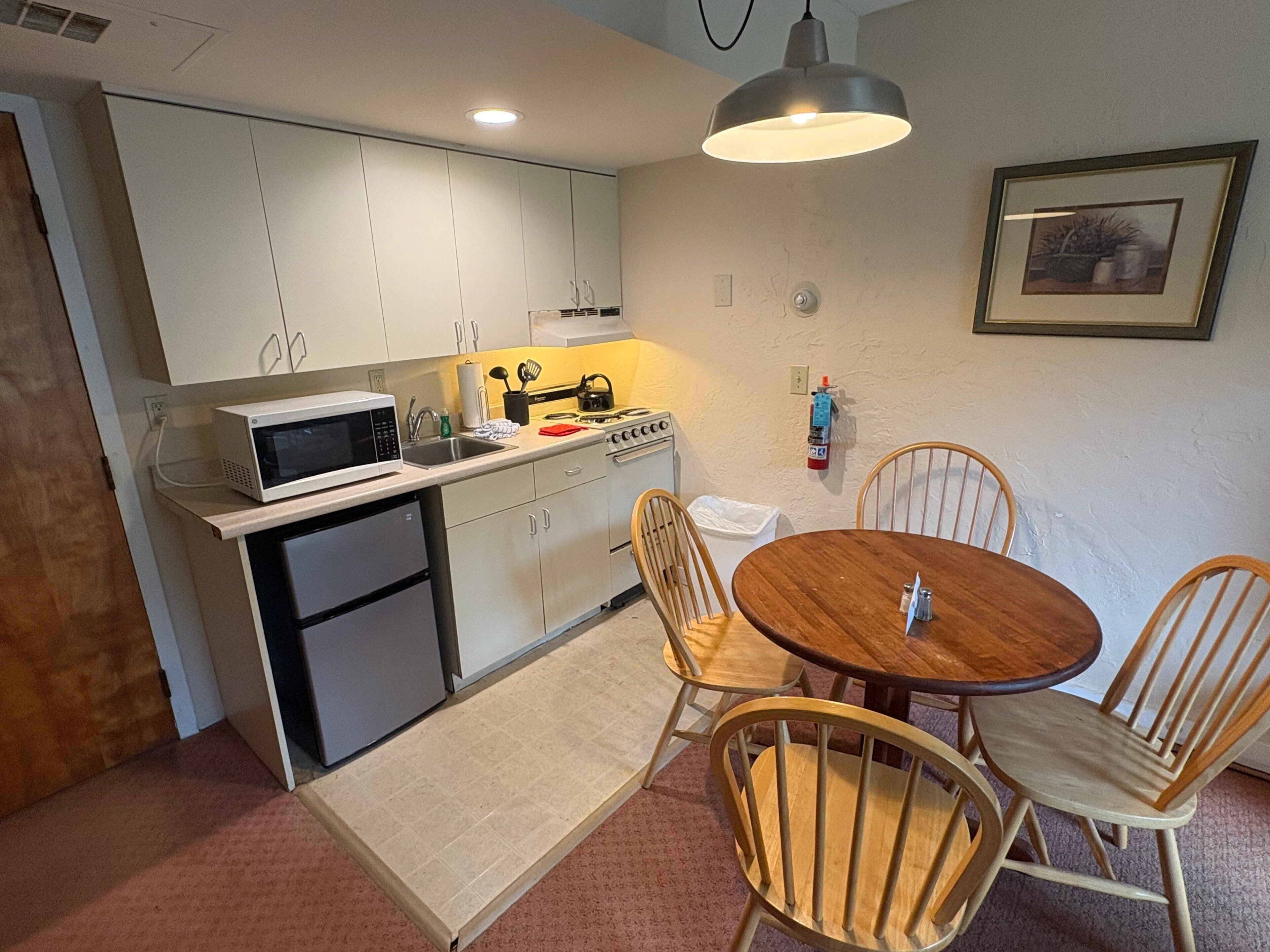 A compact kitchen with a microwave, stove, sink, and wooden chairs around a small table.