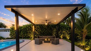 Um pátio externo com piscina, ventilador de teto e área de estar com almofadas.