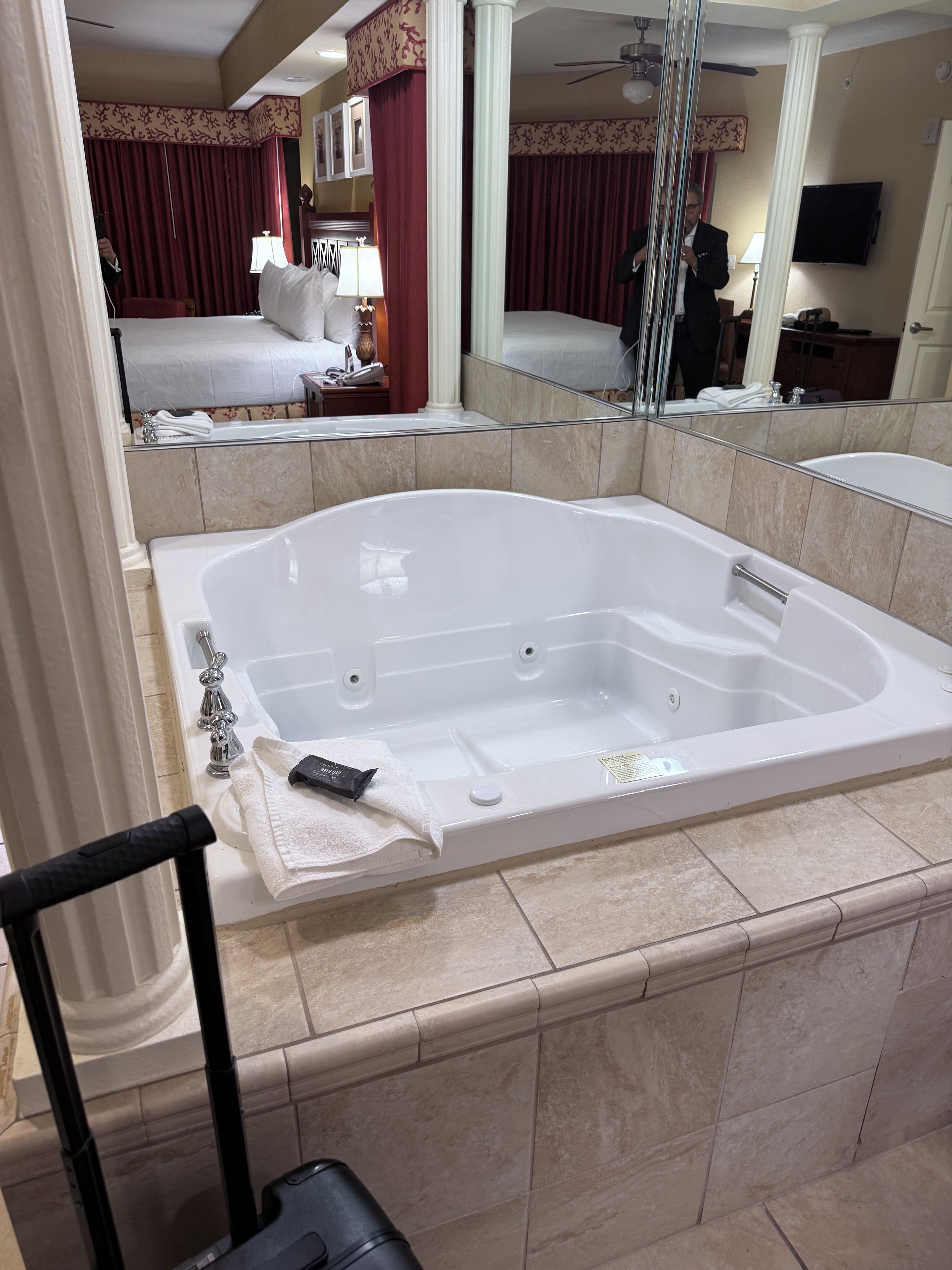 Un baño de hotel con una bañera grande, un espejo y una cama al fondo.