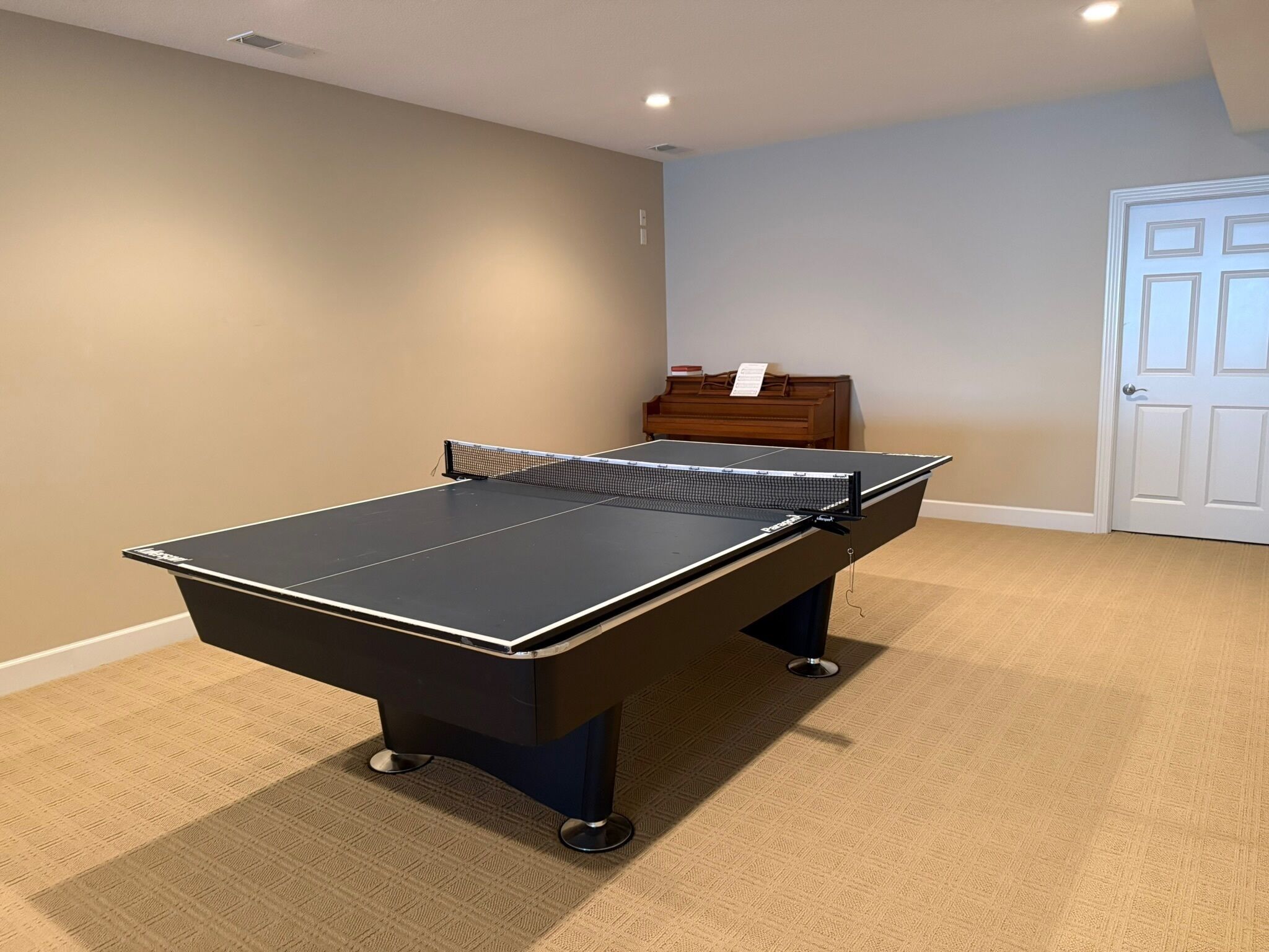 A ping pong table in a room with carpeted flooring, a piano, and a door.