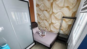 Une petite pièce comprenant un lit, une armoire, une fenêtre avec des stores et un mur décoré d’un motif.