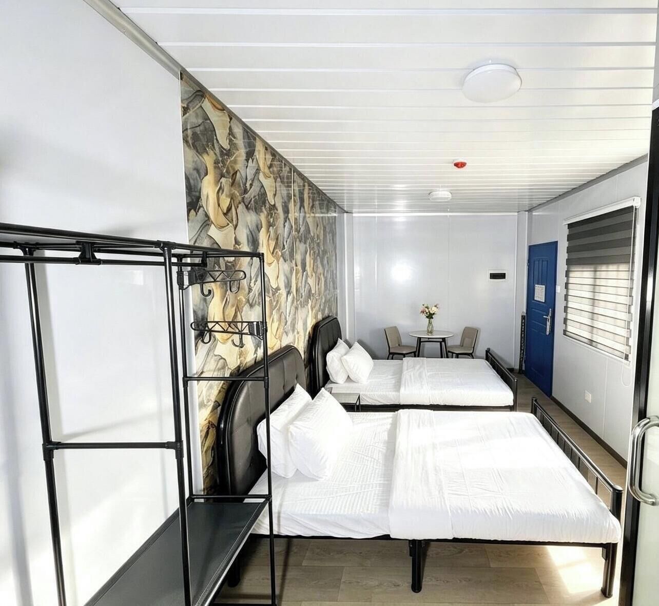 A hotel room with two beds, a small table with chairs, a blue door, and a patterned wall.