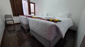 A double bed with a white duvet and a colorful blanket, a grey chair, and a window with curtains.