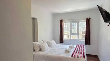 Una habitación de hotel con una cama, una mesa con un control remoto, un televisor montado en la pared y un ventanal con cortinas.