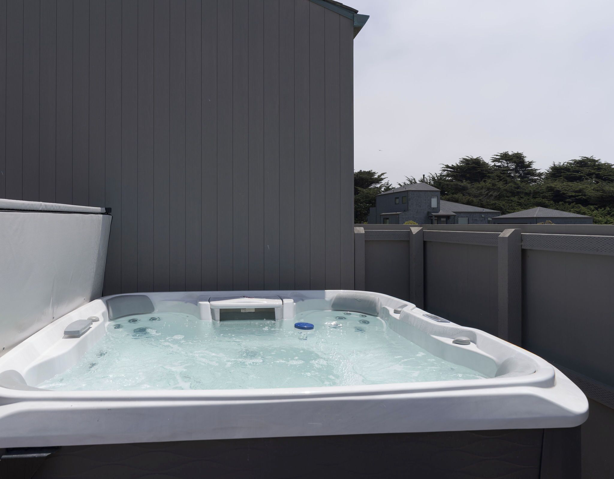 A hot tub with a control panel, surrounded by a wooden fence, with a residential area in the background.