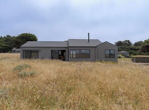 A modern two-story house with large windows, a chimney, and a surrounding field. - Driftwood Retreat | A Mindful Coastal Haven with Ocean Views & Hot Tub (The Sea Ranch)