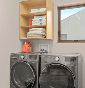 A laundry area with two front-loading washing machines, a shelf with folded towels, and a window. - Driftwood Retreat | A Mindful Coastal Haven with Ocean Views & Hot Tub (The Sea Ranch)