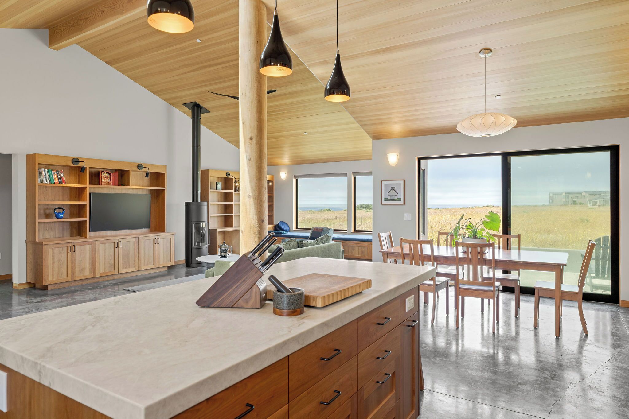 A modern kitchen with a large island, wooden cabinetry, and a view of a field through large windows.