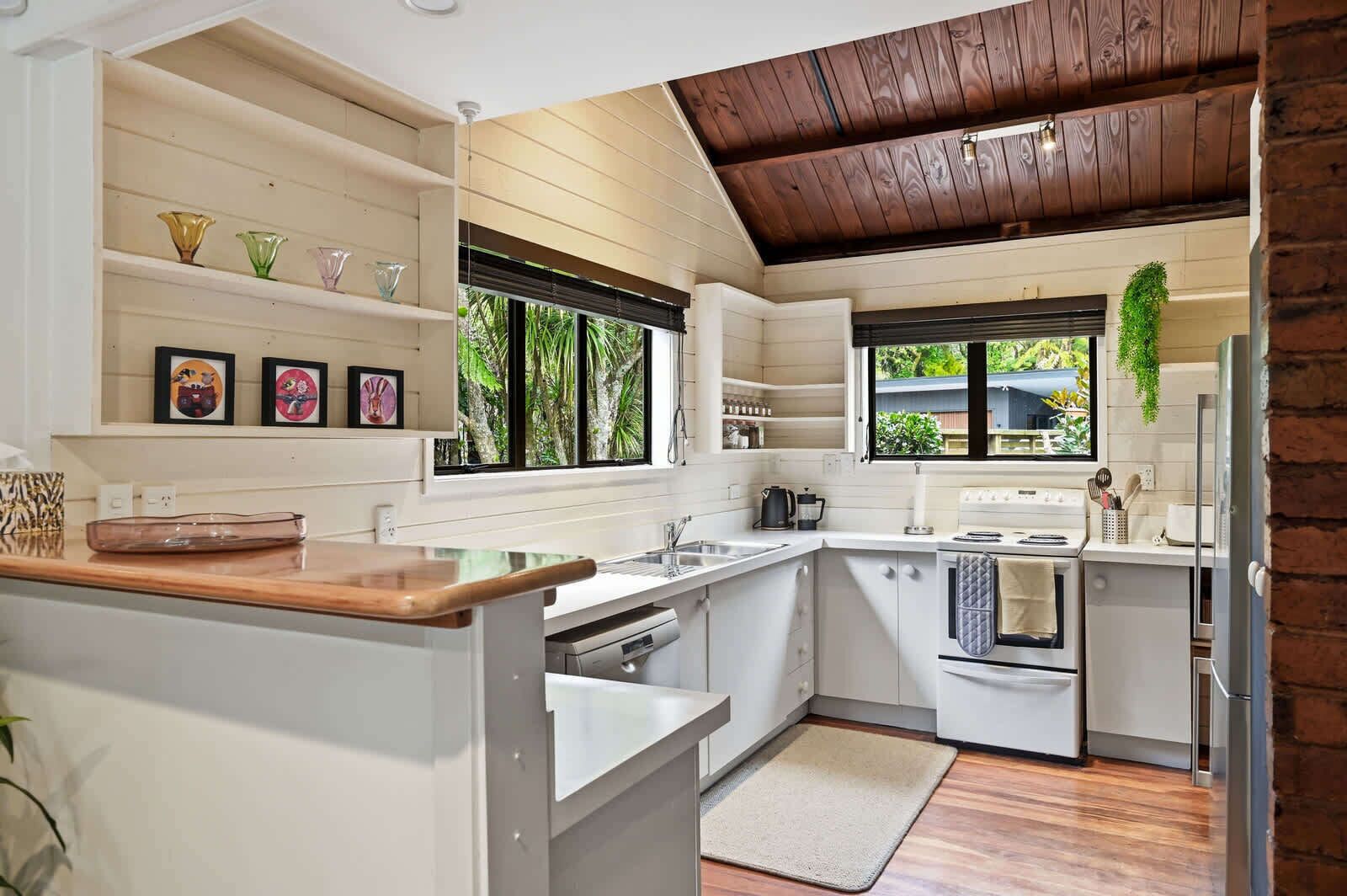 Cocina compacta con armarios blancos, encimera de madera, horno integrado y ventana con vistas a la vegetación.