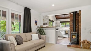 A modern living room with a beige sofa, a fireplace, and a kitchen in the background. - Tui Cottage - Walk to the Kaituna River (Okere Falls)