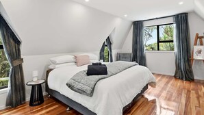 A modern bedroom with a large bed, a side table, a window with curtains, and a painting on the wall. - Tui Cottage - Walk to the Kaituna River (Okere Falls)