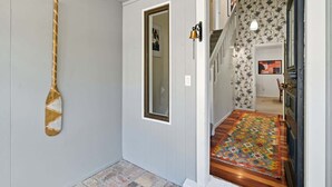 A hallway with a wooden paddle on the wall, a door with a mirror, and a patterned rug leading to a staircase. - Tui Cottage - Walk to the Kaituna River (Okere Falls)