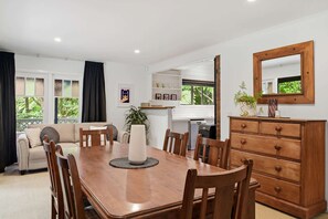 Un coin repas avec une table en bois, des chaises et un vase central. On aperçoit un canapé et une commode avec un miroir à l’arrière-plan.