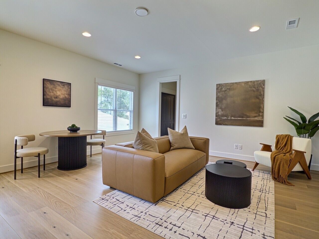 Un salon moderne avec un canapé, une table basse et des fauteuils. On y trouve une fenêtre avec des stores, une porte et un tableau accroché au mur.