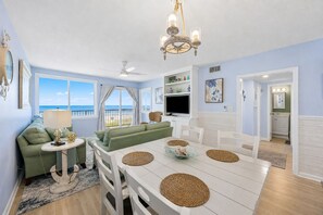 A living room with a sofa, a dining table with placemats, a television, and a view of the ocean. - Ocean-view 2BR condo in North End Fernandina Beach. (Fernandina Beach)