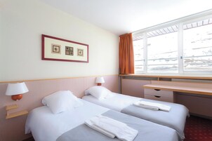 Et hotelværelse med to senge, et skrivebord, et vindue med gardiner og et billede i ramme på væggen.