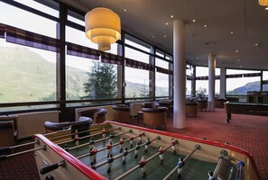 Une pièce spacieuse équipée d’un baby-foot, de sièges confortables et de grandes fenêtres offrant une vue sur l’extérieur.