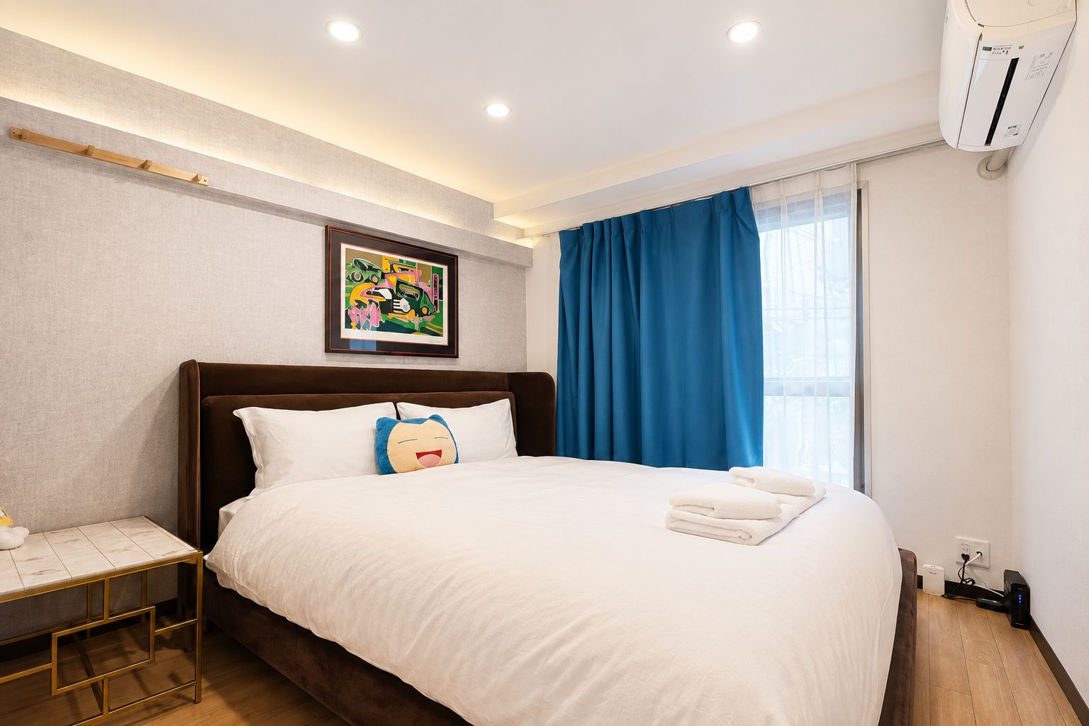 A hotel room with a large bed, a bedside table, a wall-mounted air conditioner, and a window with blue curtains.
