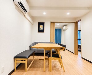 A room with a wooden table, bench, and grey sofa. There is a framed picture on the wall and an air conditioning unit mounted on the wall. - SERENE | Nishikujo, 4th Floor | 60 ㎡, Sleeps 7 | Direct Access to USJ and Osaka Station (大阪市)