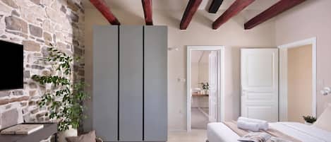 Un dormitorio con pared de piedra, una cama con ropa blanca, un armario gris y un televisor a la izquierda.