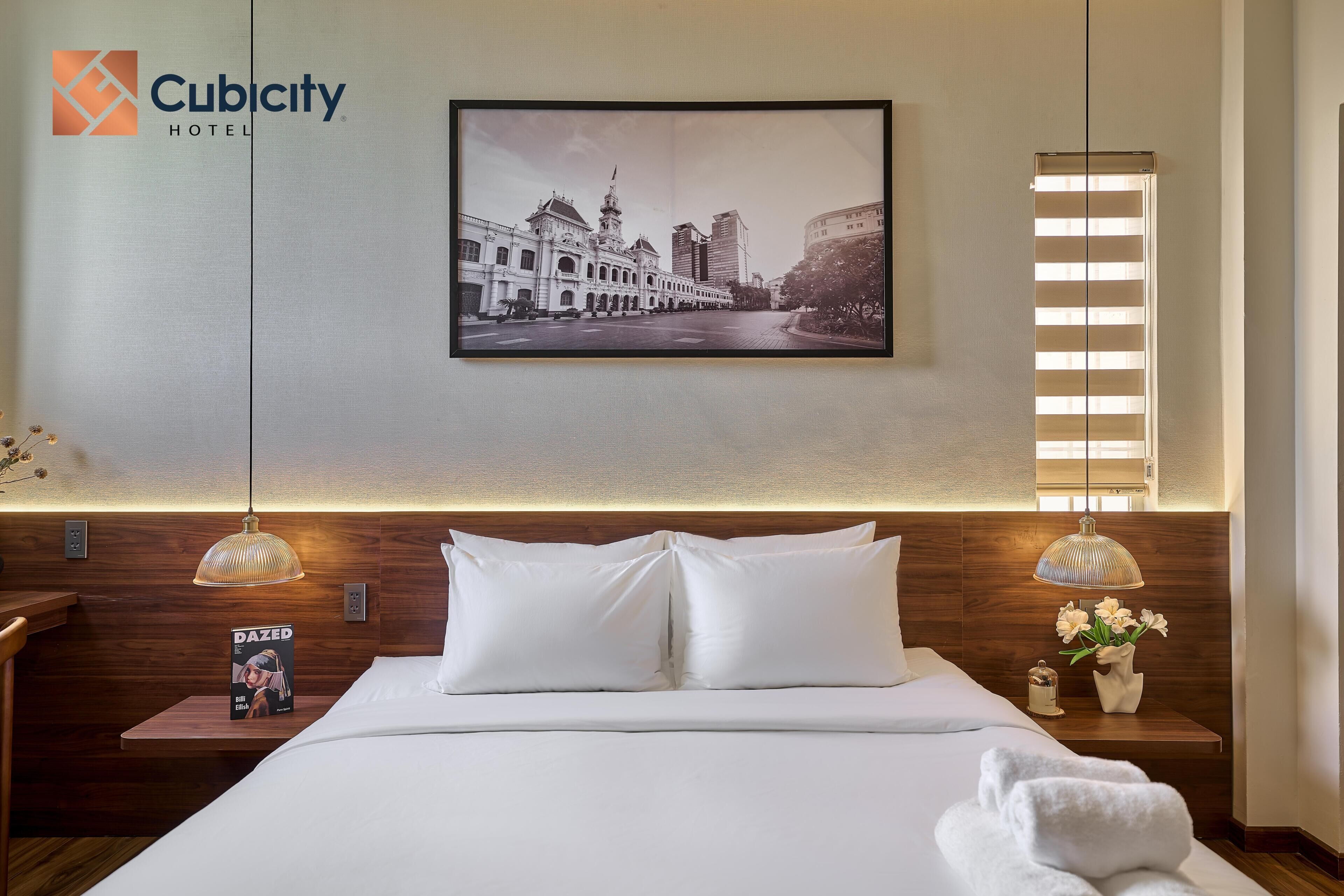 A hotel room with a large bed, two bedside lamps, a framed picture on the wall, and a window with blinds.