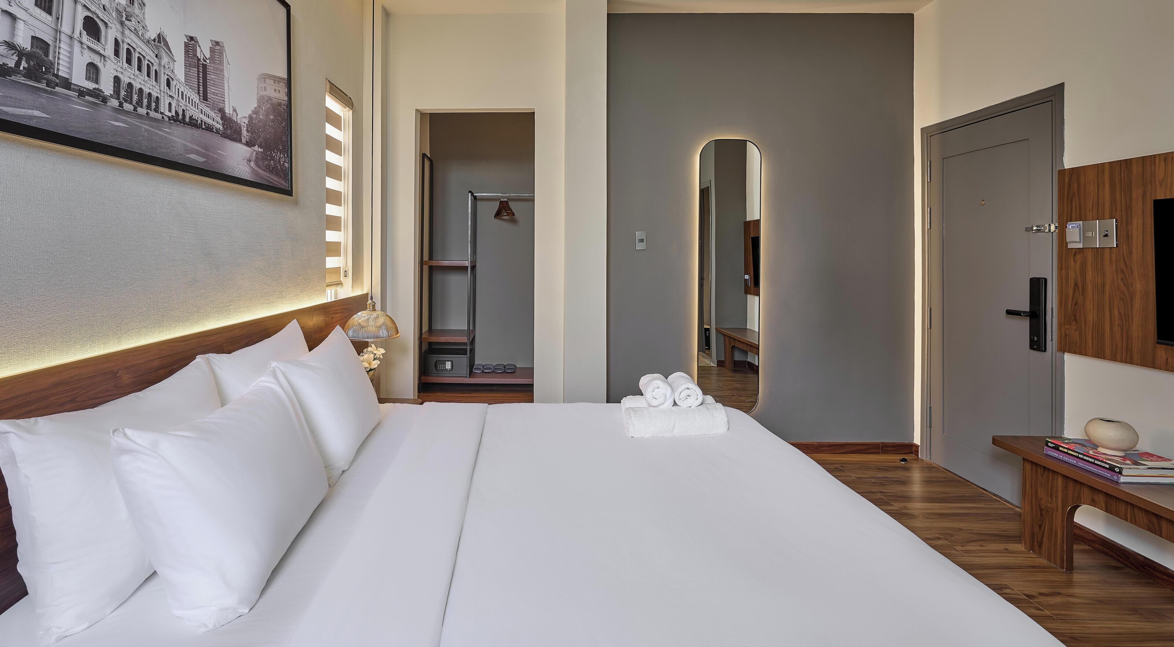 Habitación de hotel con una cama grande, cabecera de madera, mesita de noche con lámpara, un espejo y vistas a la ciudad desde la ventana.