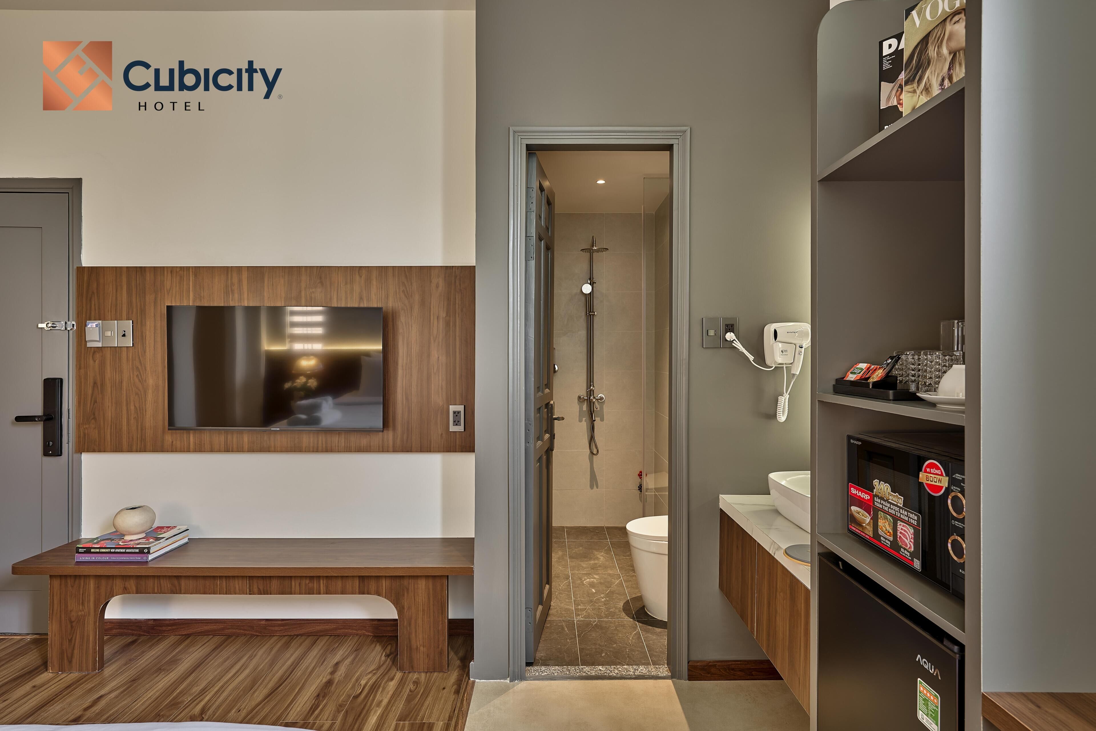 A hotel room with a wooden bench, a TV mounted on the wall, a shower area, and a minibar.