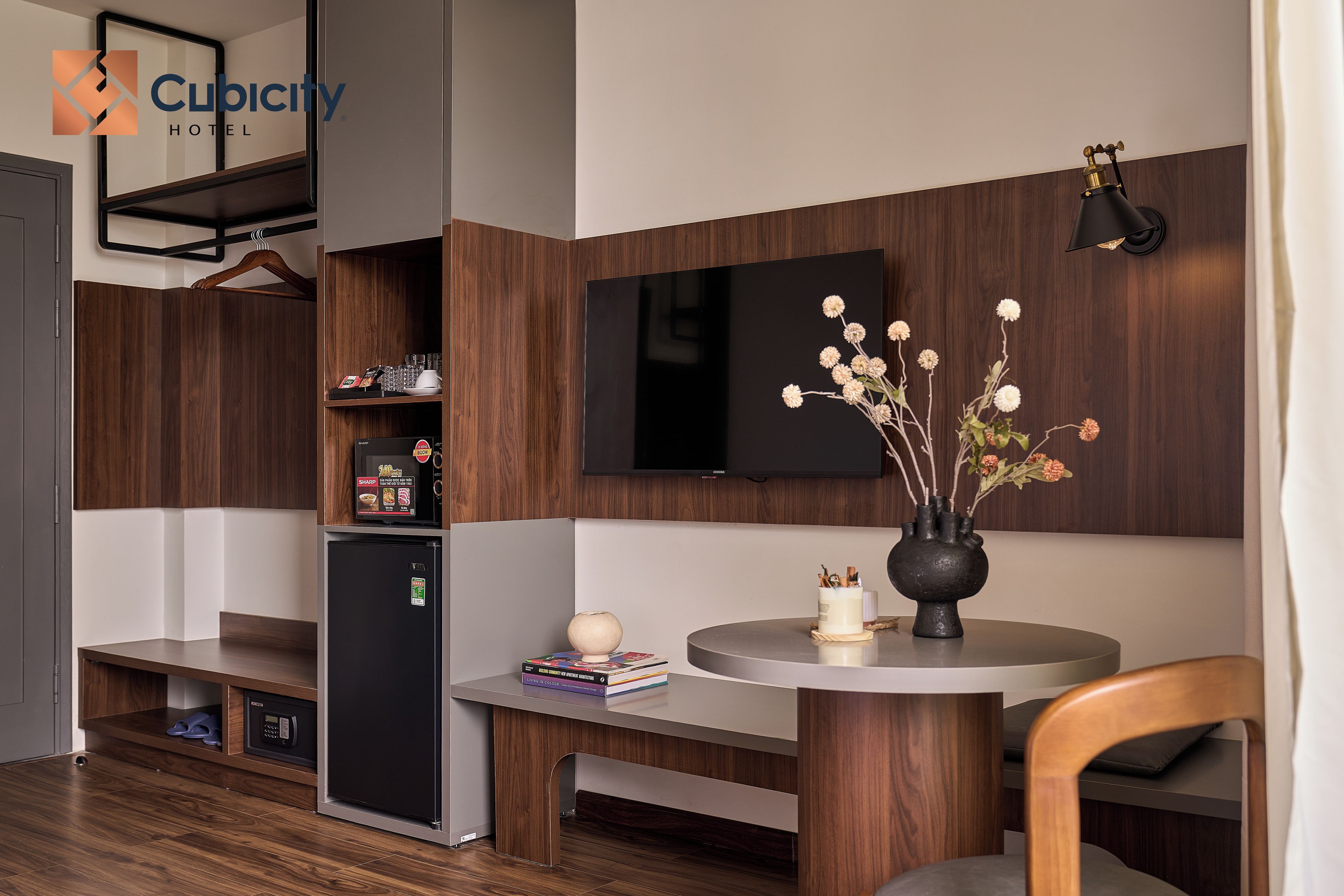 Ein Hotelzimmer mit einem Holzkasten, einem schwarzen Kühlschrank, einem Flachbildfernseher, einem runden Tisch mit Blumenvase und einer Pendelleuchte.