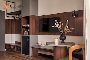 Una habitación de hotel con un armario de madera, un refrigerador negro, un televisor de pantalla plana, una mesa redonda con un jarrón de flores y una lámpara colgante.