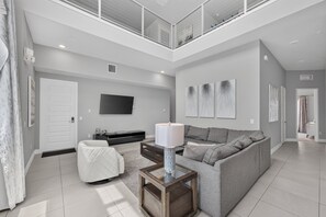 A modern living room with a high ceiling, a flat-screen TV mounted on the wall, a grey sofa, a coffee table, and a white chair. - Reunion Resort 5BR Condo | Park View | Resort Pool (Kissimmee)